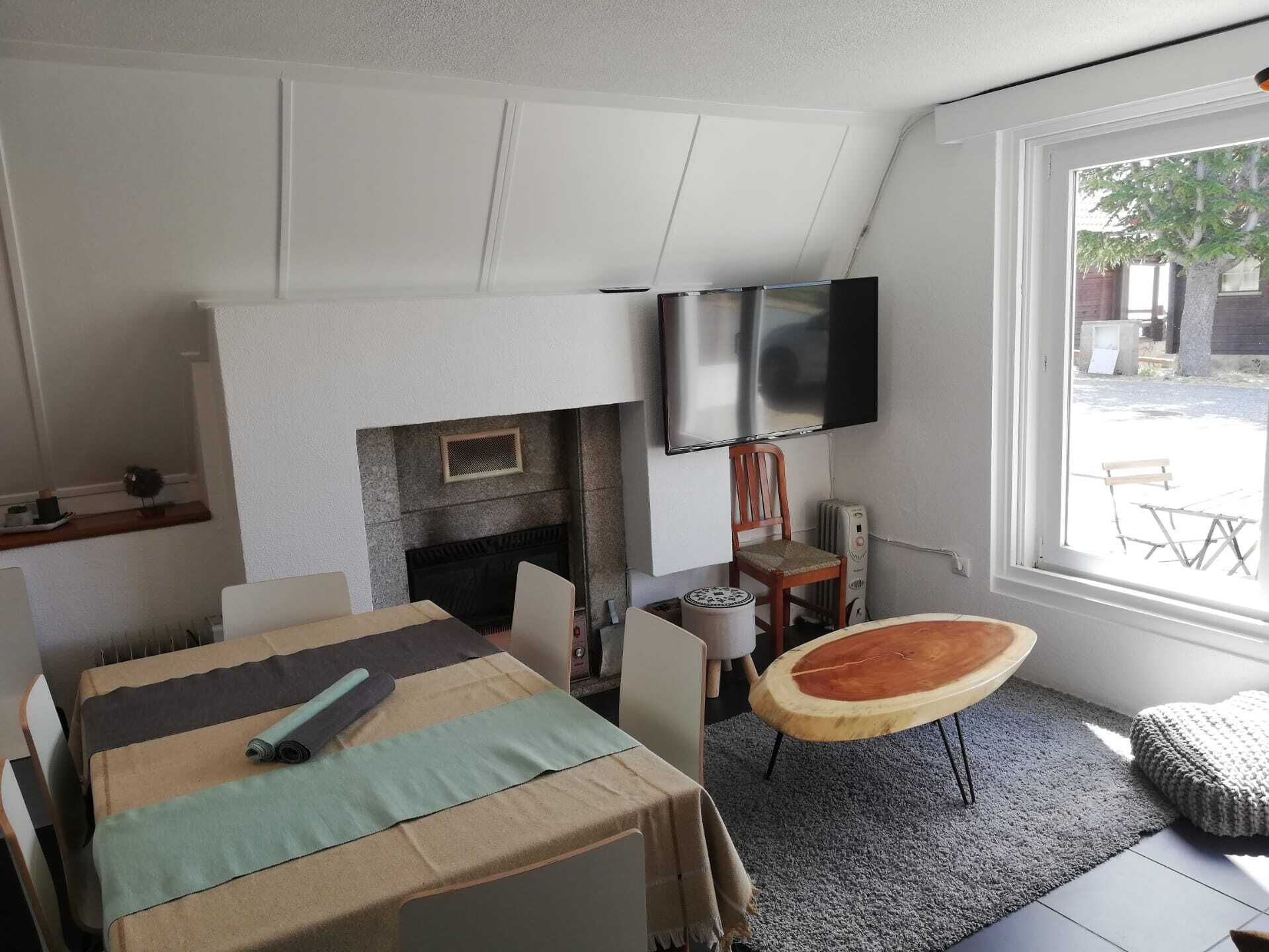 A room with a fireplace, a flat-screen TV, a dining table with chairs, a wooden chair, and a window with a view of the outside.