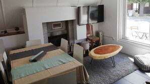 Ett rum med en öppen spis, en platt-TV, ett matbord med stolar, en trästol och ett fönster med utsikt över omgivningen.