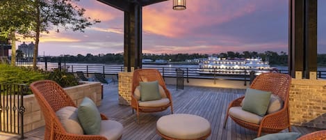 A covered outdoor seating area with wicker chairs and a wooden deck, overlooking a river with a boat and cityscape in the background.