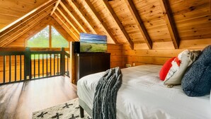 Una camera da letto con soffitto in legno, una televisione su un mobile, un letto con cuscini e vista su un paesaggio verde attraverso una finestra.