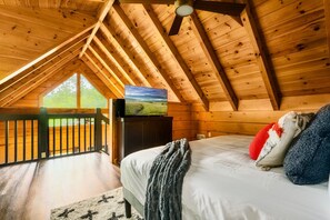 A bedroom with a wooden ceiling, a television on a cabinet, a bed with pillows, and a view of a green landscape through a window. - 5* IndoorPool~Fireplace~FireTbl~Hot Tub~PS5~Bears (Gatlinburg)