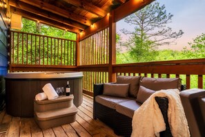 A wooden deck with a hot tub, wicker sofa, and a view of greenery. - 5* IndoorPool~Fireplace~FireTbl~Hot Tub~PS5~Bears (Gatlinburg)