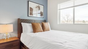 A neatly made bed with white and brown pillows, a wooden headboard, a bedside table with a lamp, and a framed abstract painting on the wall. - Landing | Amazing 2BD in Harvey Park South (Englewood)