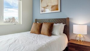 A neatly made bed with white linens and two brown pillows, a wooden headboard, a bedside lamp, and a framed abstract artwork on the wall. - Landing | Amazing 2BD in Harvey Park South (Englewood)