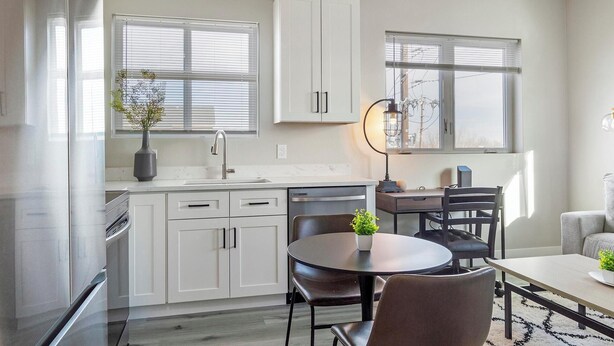 A modern kitchen with white cabinets, a dining area with a round table and chairs, and a window with blinds. - Landing | Amazing 2BD in Harvey Park South (Englewood)
