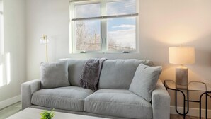 A grey sofa with a throw blanket, a small table with a lamp, and a window with blinds. - Landing | Amazing 2BD in Harvey Park South (Englewood)