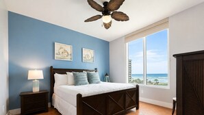Ein Schlafzimmer mit Bett, Nachttisch, Lampe, Deckenventilator und einem großen Fenster mit Meerblick.