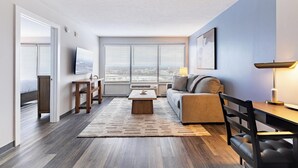 Uma sala de estar moderna com sofá, mesa de centro e TV. Há uma janela com persianas, uma porta que dá para um quarto e vista para a cidade.