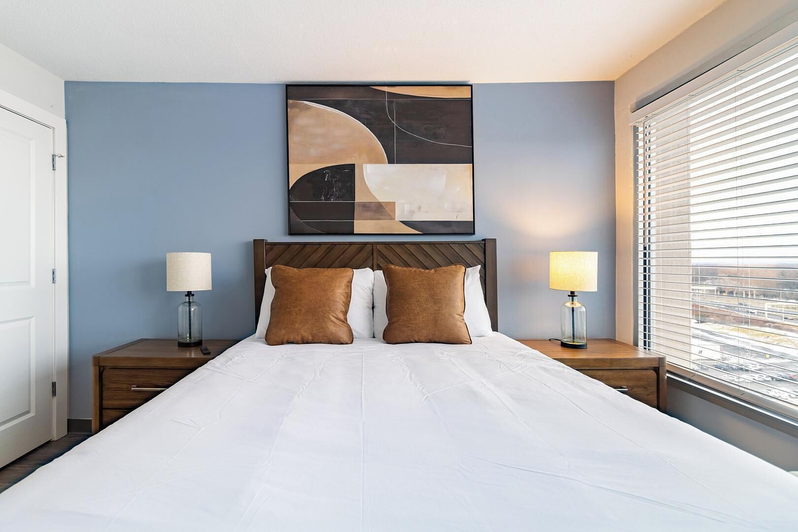 Ein Hotelzimmer mit einem großen Bett, zwei Nachttischen, einem Wandbild und einem Fenster mit Jalousien.