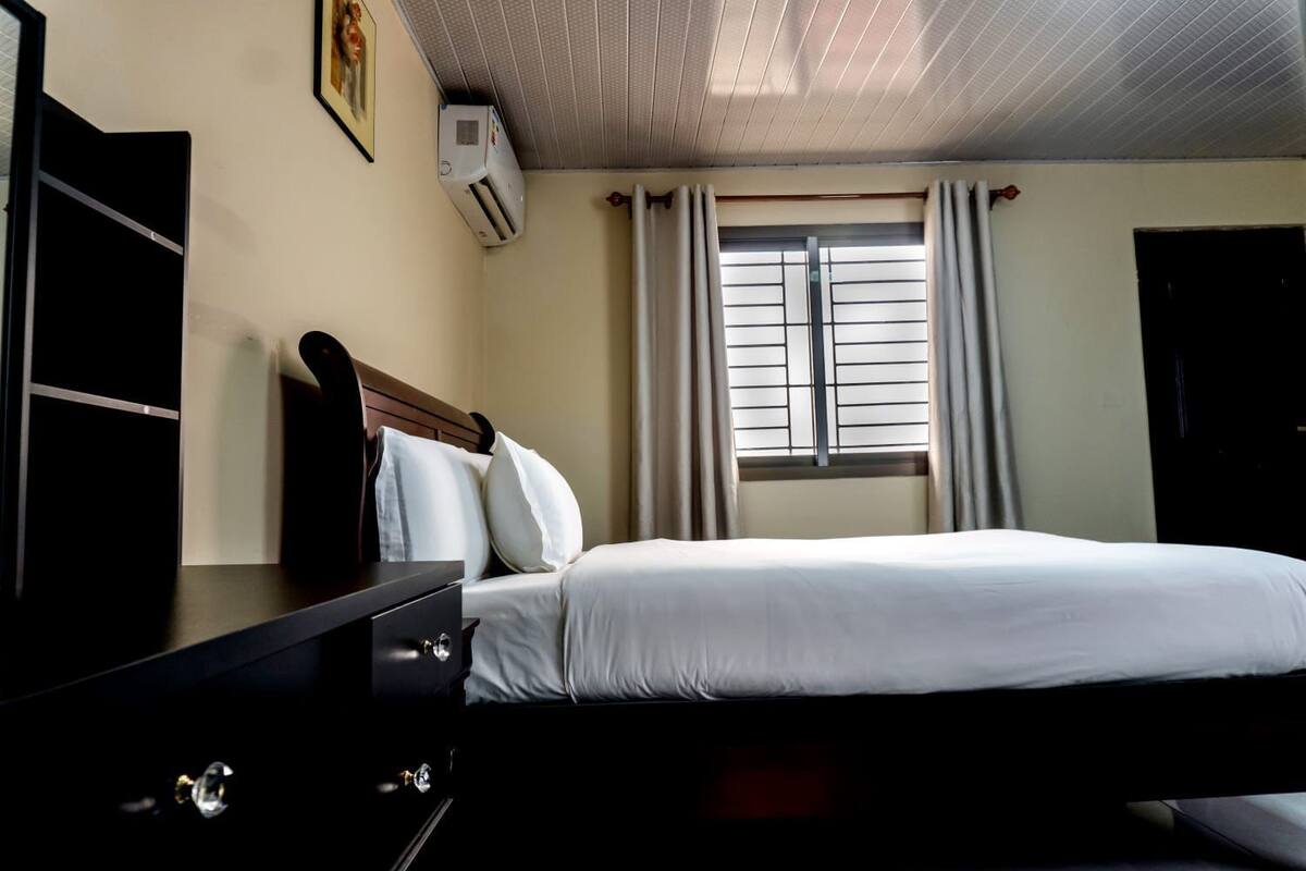 Habitación de hotel con cama, cómoda, unidad de aire acondicionado, ventana con cortinas y puerta.