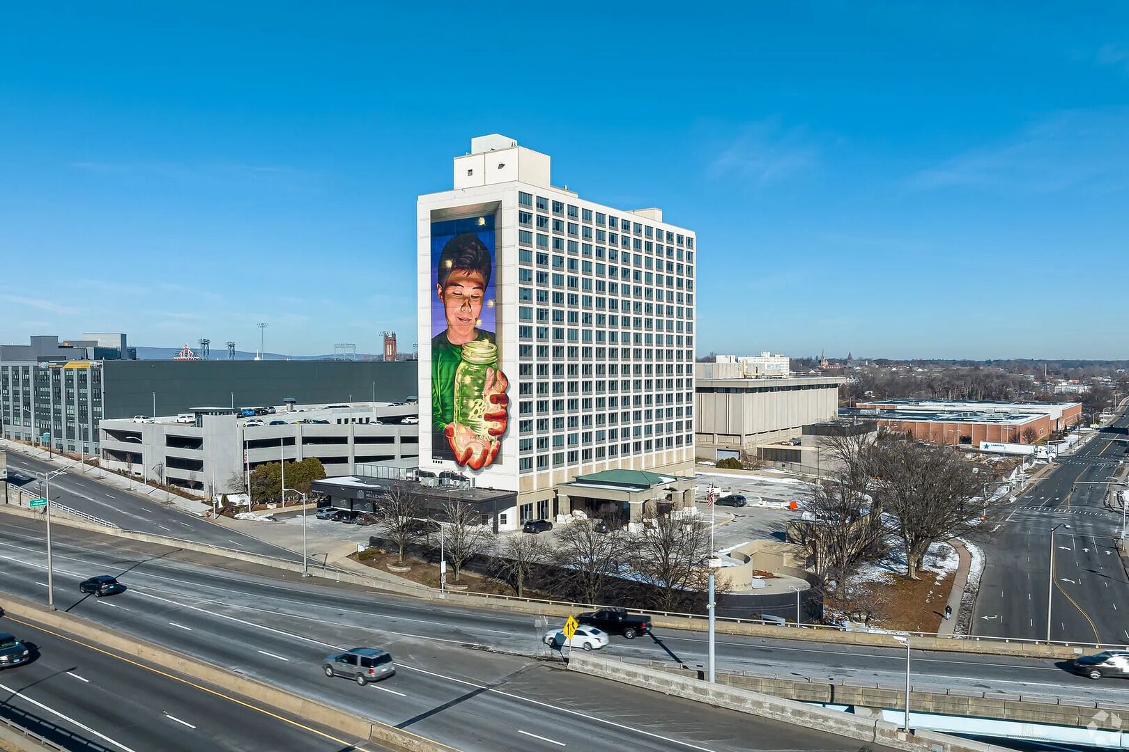 Un edificio de gran altura con un mural grande en la fachada, un estacionamiento y una autopista al frente.