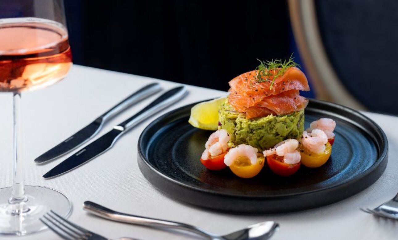 A dish with avocado, shrimp, and salmon on a plate, accompanied by a glass of wine and cutlery.