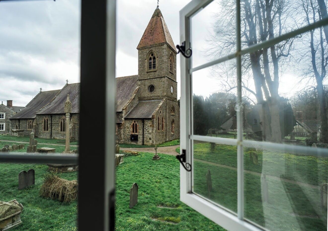 A view through a window of a church with a prominent tower, a grassy cemetery, and stone buildings.