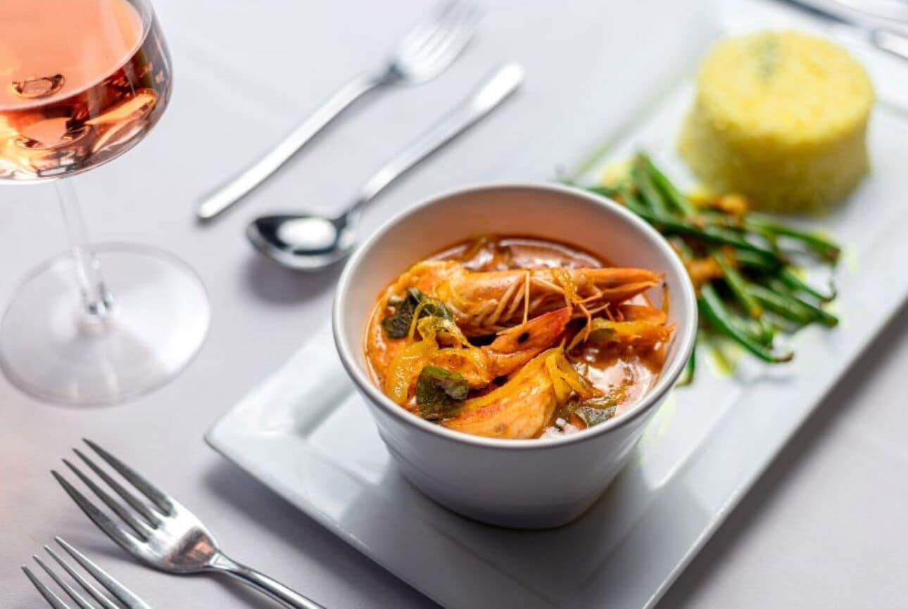 Un bol de soupe de fruits de mer aux légumes, un verre de vin rosé et une assiette avec un aliment jaune et des légumes verts.