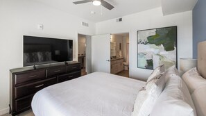 A bedroom with a large bed, a TV on a dresser, a ceiling fan, and a bathroom visible through an open door. - Landing | Incredible 2BD, Gym, Clubhouse (Mesa)