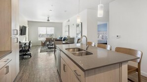 Een moderne keuken met een centraal eiland, een zithoek met een tafel en stoelen, en een slaapkamer met een bed en raam.