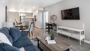 Een moderne woonkamer met een bank, salontafel en tv-meubel. Achtergrond is een keuken met kastjes en een eethoek.