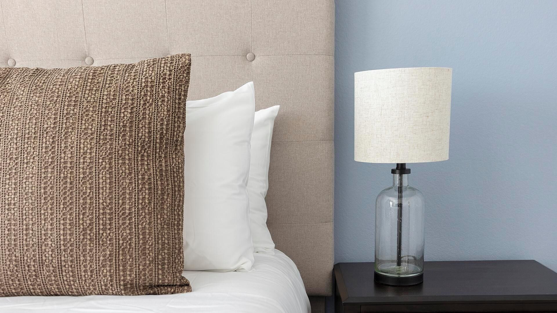A bed with a tufted headboard, a brown textured pillow, a white pillow, and a clear glass lamp on a dark nightstand.