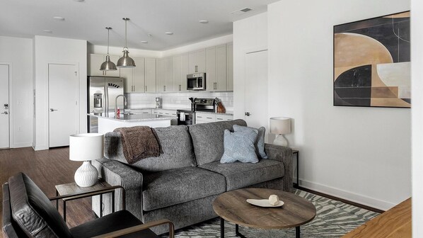 A modern living room with a grey sofa, a coffee table, and a dining area with a kitchen in the background. - Landing | Spectacular 2BD, Clubhouse, Gym (Raleigh)