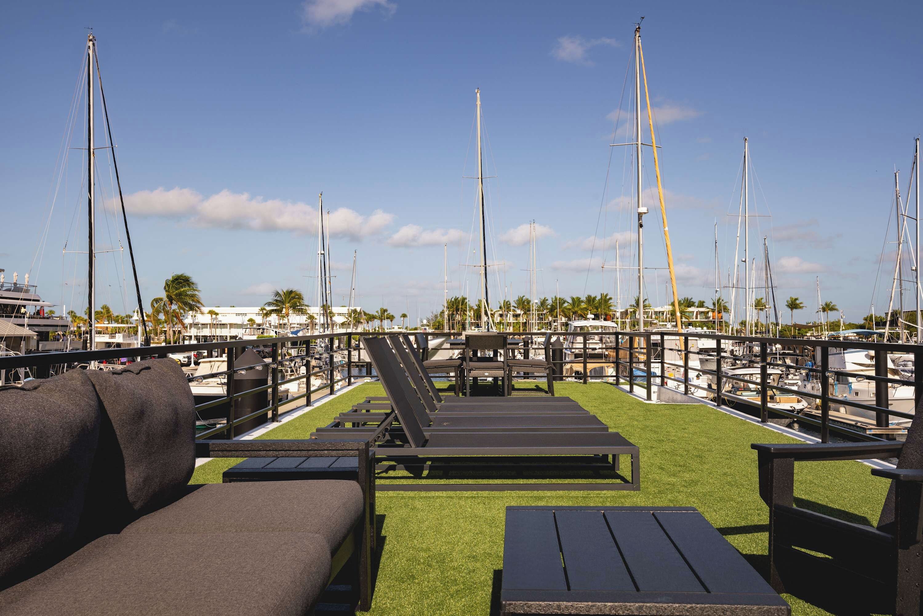 A marina with multiple sailboats docked, a grassy deck with lounge chairs, and a clear blue sky.