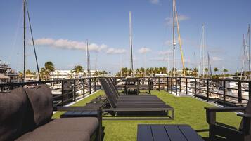 A marina with multiple sailboats docked, a grassy deck with lounge chairs, and a clear blue sky.