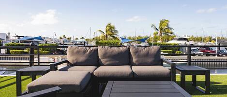 A waterfront outdoor seating area with a sofa and tables, overlooking a marina with boats and palm trees.