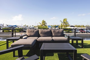 A waterfront outdoor seating area with a sofa and tables, overlooking a marina with boats and palm trees. - Waterfront & Heated Pool - Break Awai Floating Villa (Stock Island)
