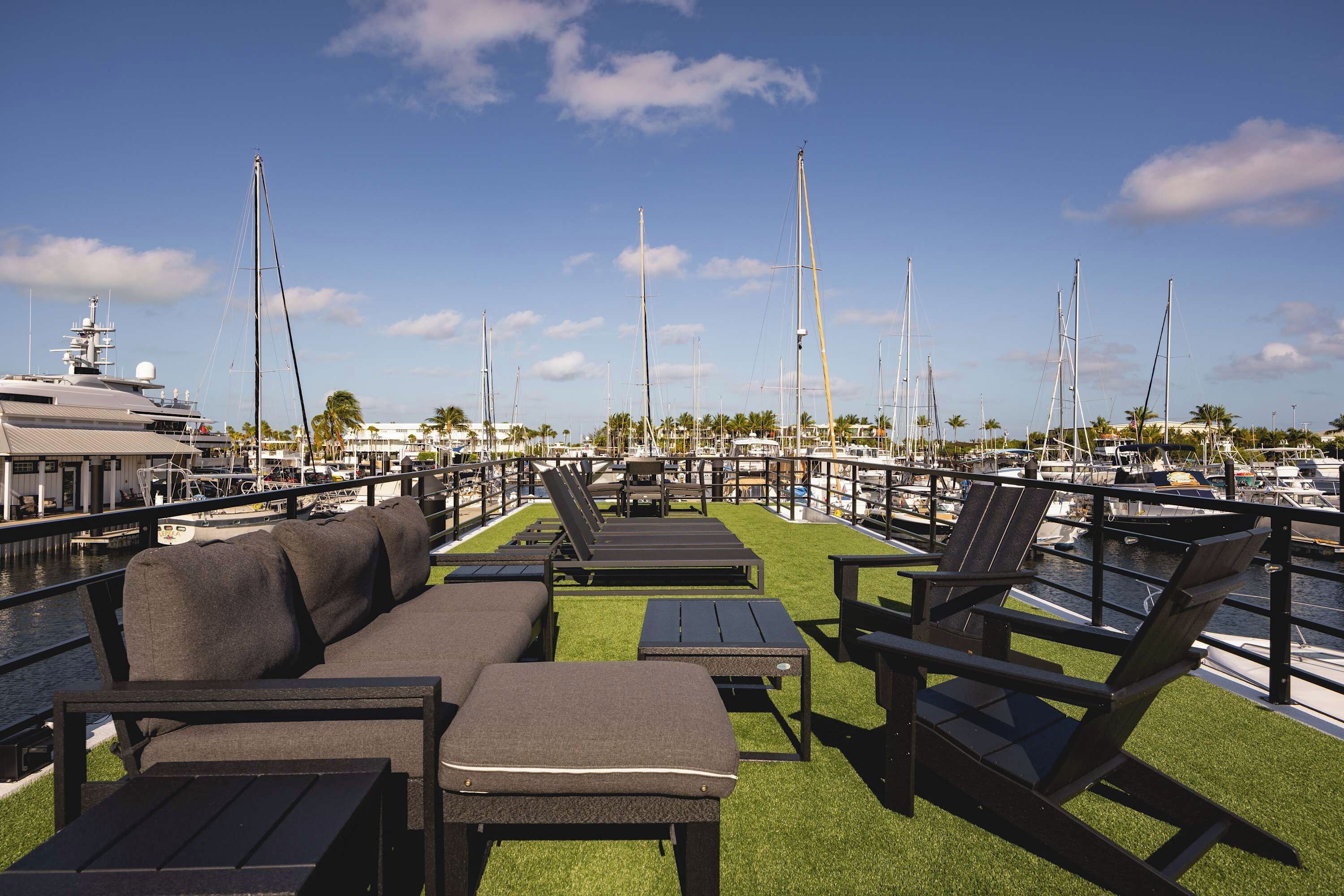 A marina with a row of lounge chairs and a small table on a grassy deck, overlooking a marina with several sailboats.