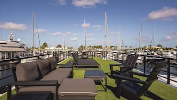 A marina with a row of lounge chairs and a small table on a grassy deck, overlooking a marina with several sailboats.