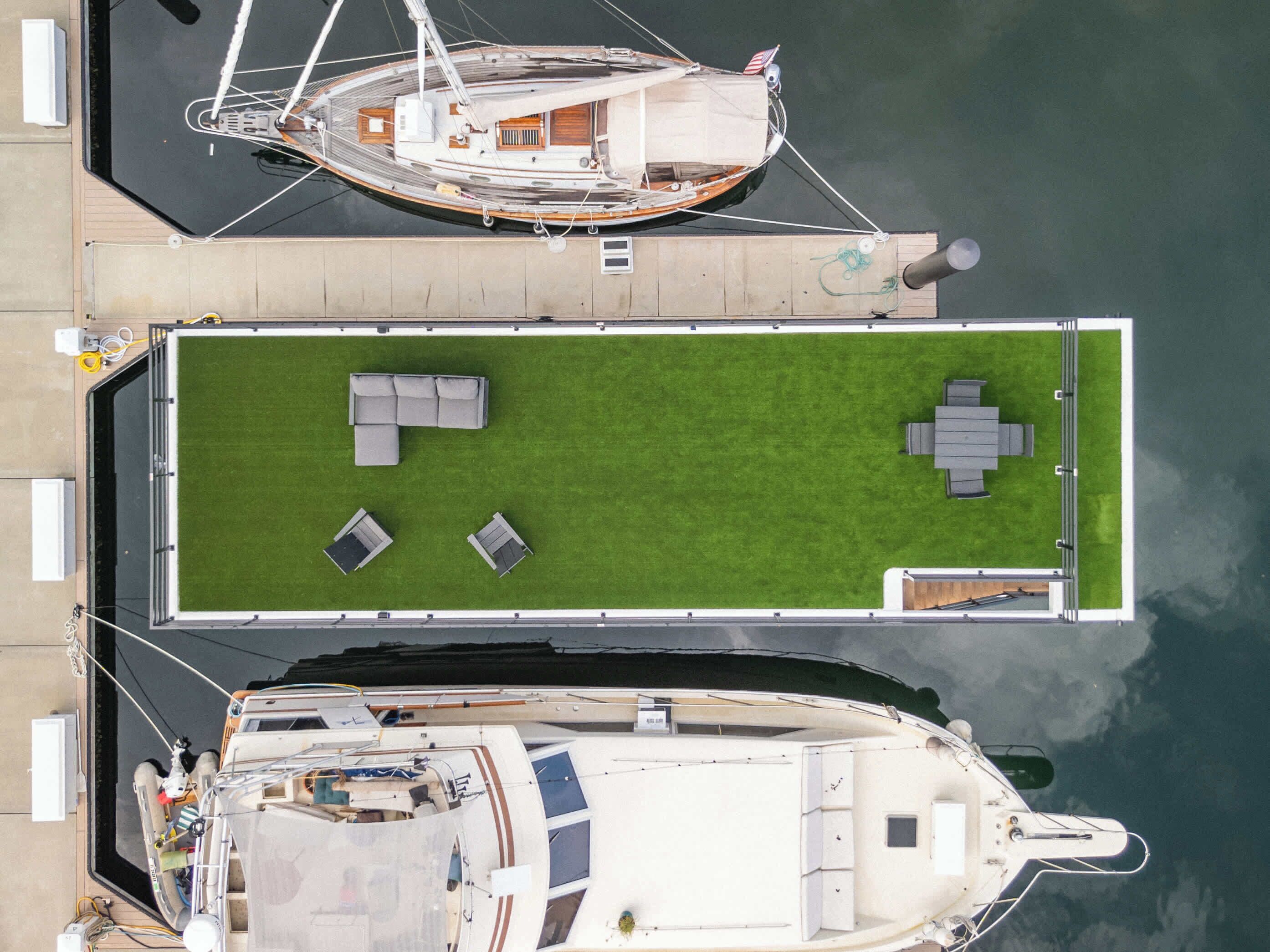 Un porto turistico con due imbarcazioni ormeggiate, una zona prendisole con mobili da esterno e un piccolo edificio.