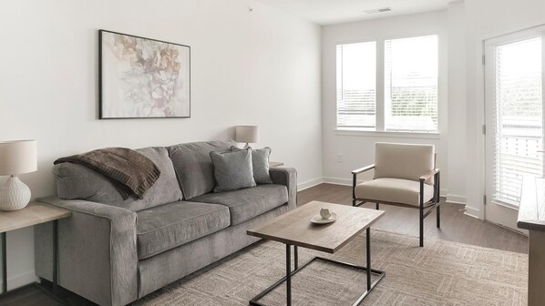 Een moderne woonkamer met een grijze bank, een klein tafeltje, een stoel en een raam met zonwering.