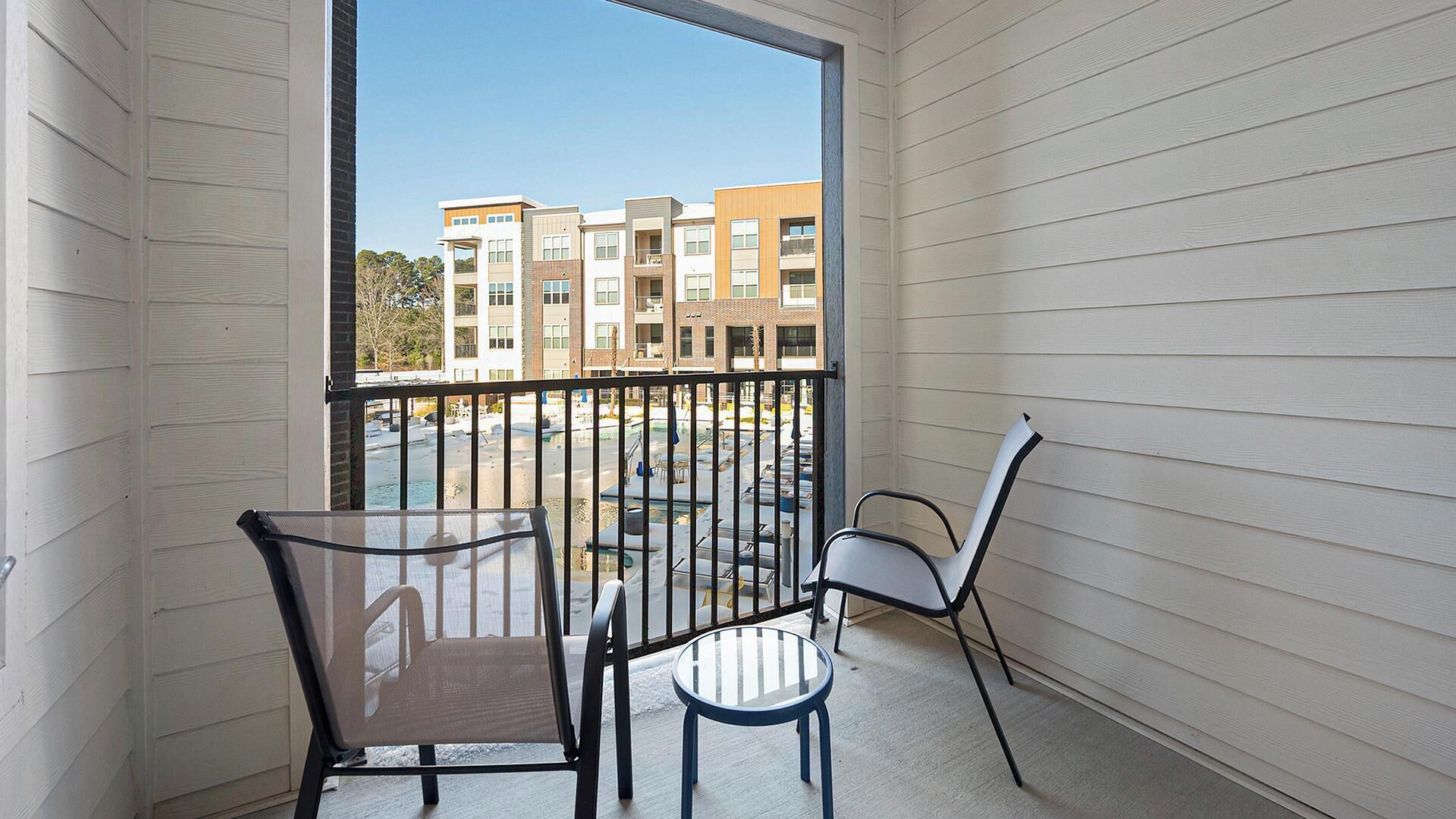 A balcony with two chairs and a small table, overlooking a parking lot and multi-story buildings.
