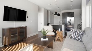 A modern living room with a flat-screen TV, a dark wooden cabinet, a sofa with patterned cushions, and a dining area with a wooden table and a potted plant.