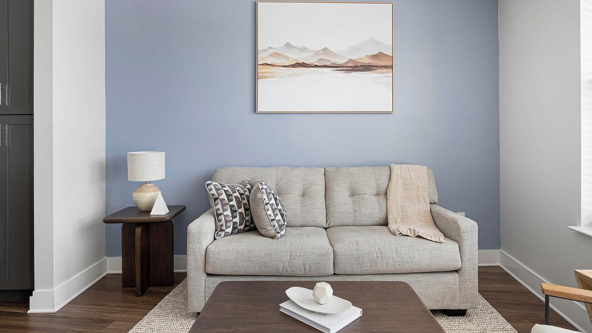 Ein Wohnzimmer mit grauem Sofa, einem kleinen Beistelltisch mit Lampe und einem gerahmten Landschaftsbild an der Wand.