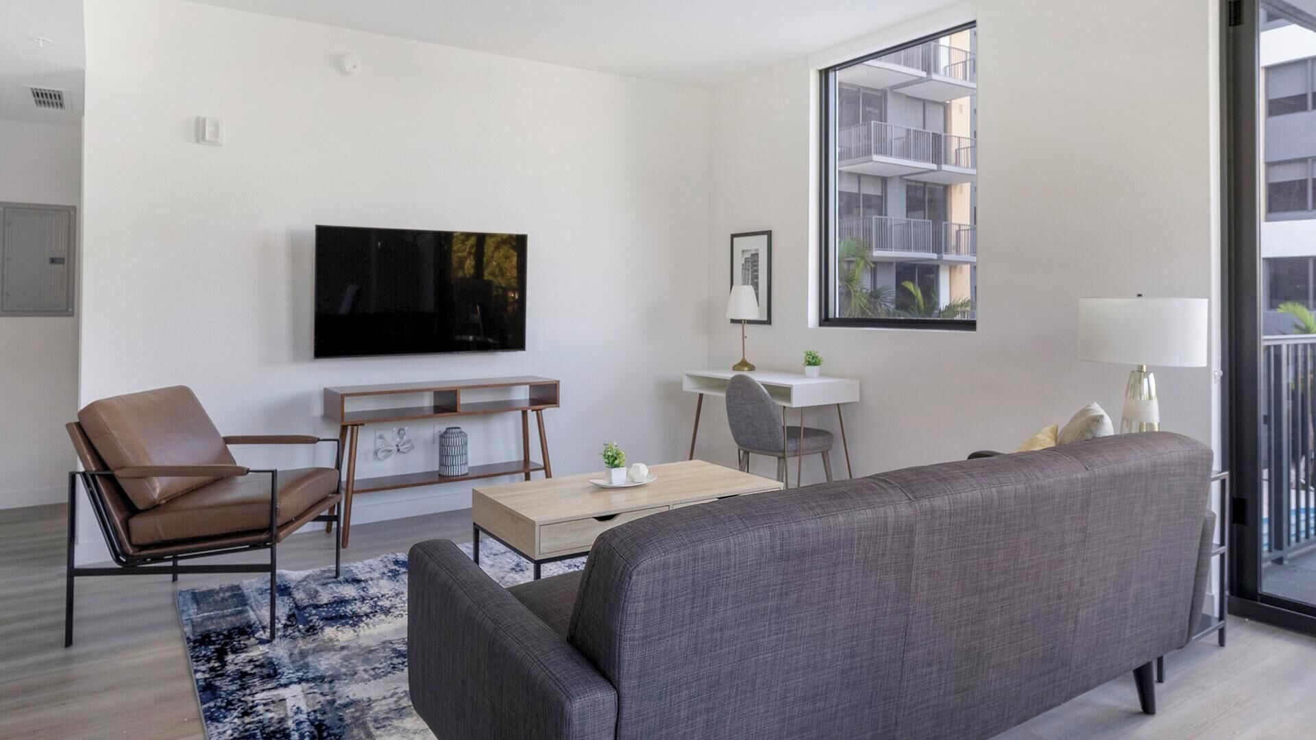 A modern living room with a sofa, a chair, a coffee table, a TV, and a balcony with a view of buildings.