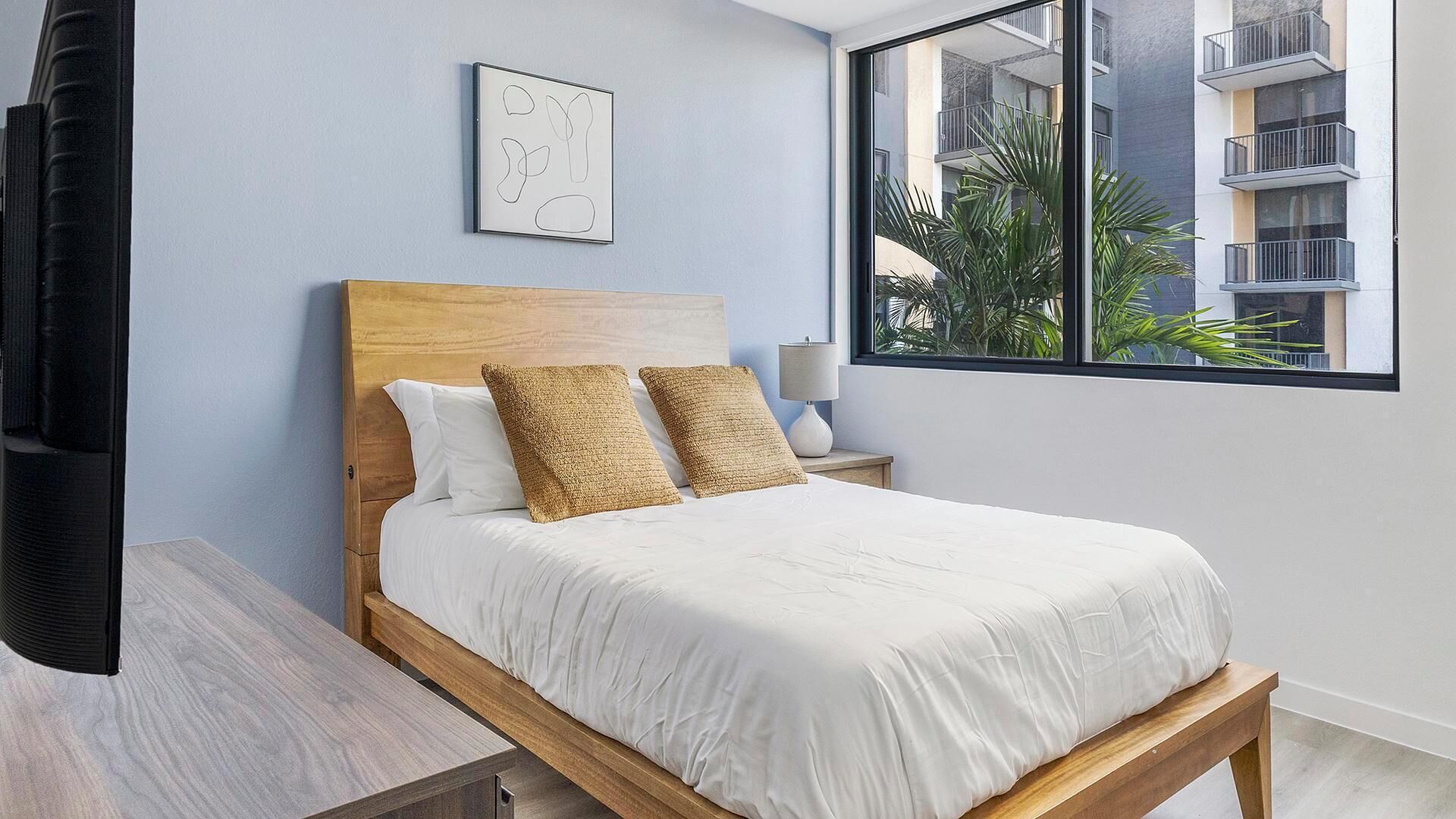 A bedroom with a wooden bed, white bedding, a bedside lamp, a framed abstract artwork, and a window with a view of buildings and palm trees.