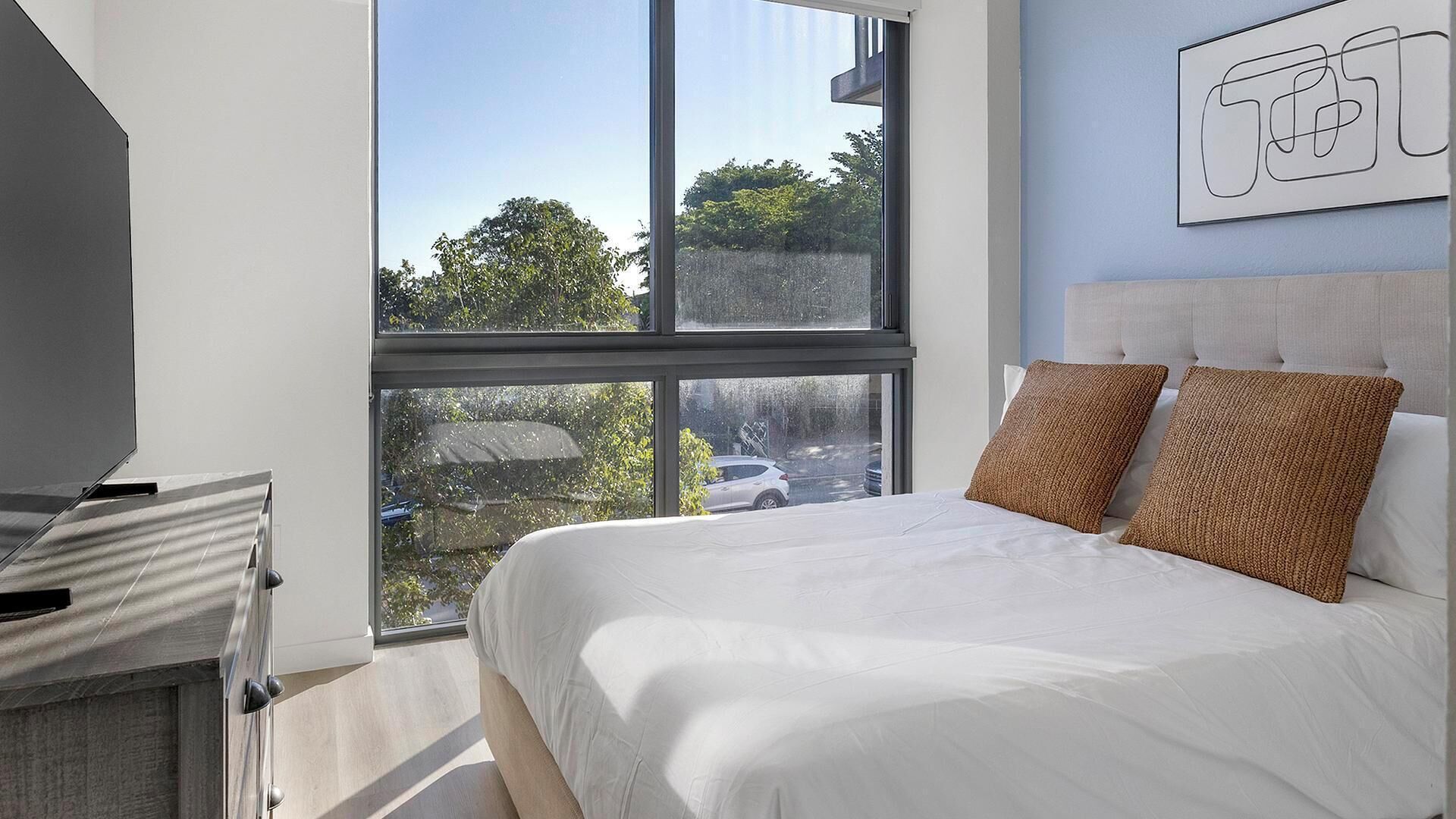 A modern bedroom with a large window, a bed with white bedding and two brown pillows, and a wall-mounted artwork.