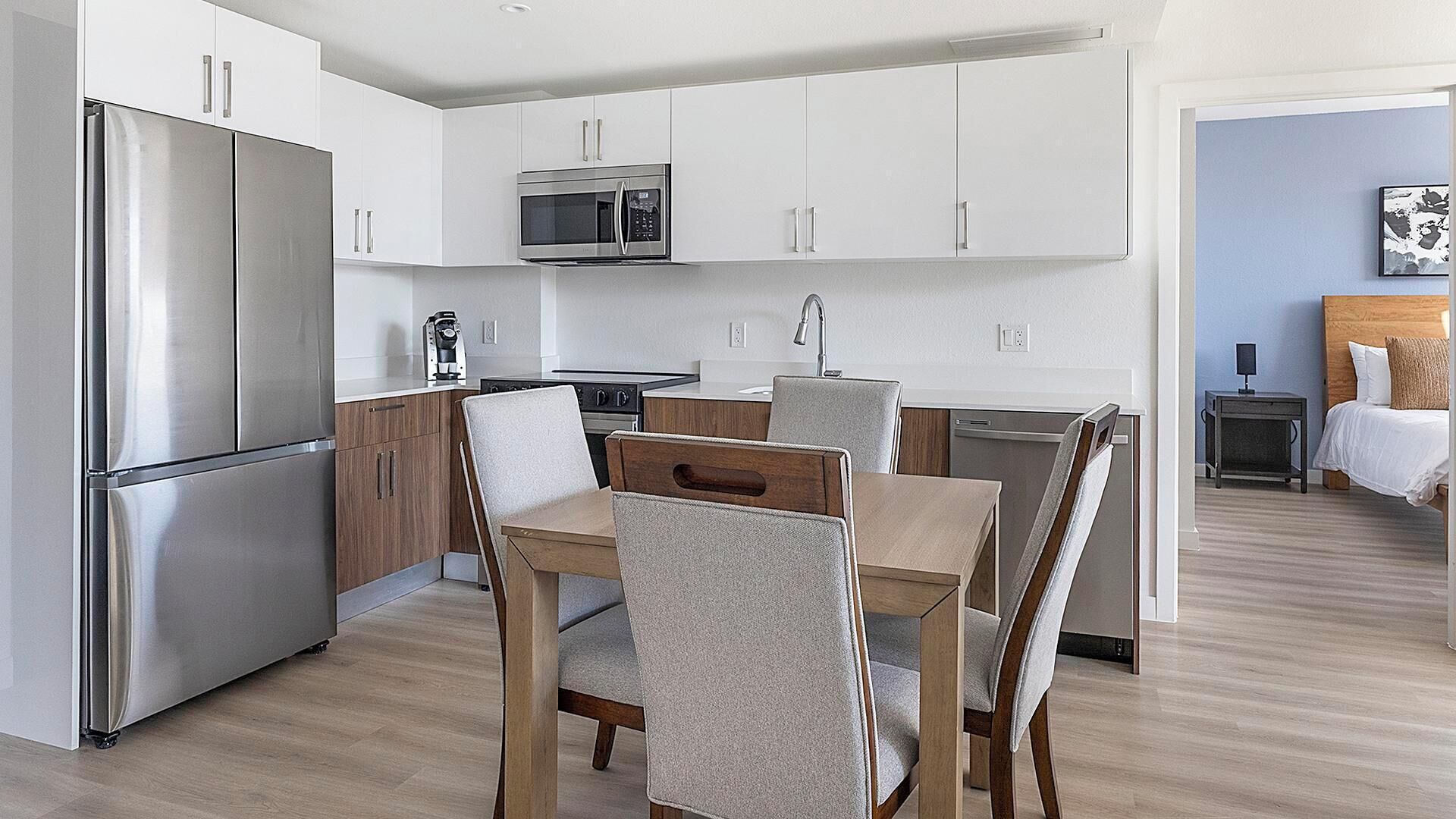 A modern kitchen with stainless steel appliances, a dining area with a wooden table and chairs, and a bedroom with a bed and nightstand.
