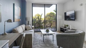 A modern living room with a sofa, coffee table, and chairs, a flat-screen TV, and a balcony with plants. - Landing | Stunning 1BD, Yoga Studio, Game Room (Miami)