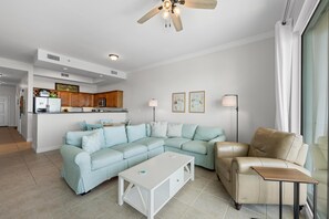 A living room with a sectional sofa, a coffee table, a leather armchair, and a dining area with wooden cabinets and a refrigerator. - The Indies 609 (Gulf Shores)