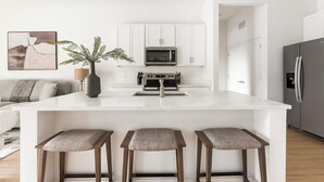 A modern kitchen with white cabinetry, a central island with stools, a microwave, and a refrigerator. - Landing | Beautiful 2BD, Pool (Casas Adobes)