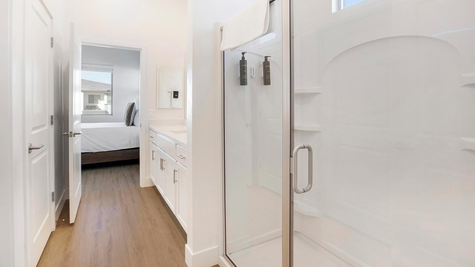Un couloir étroit avec un lit, une salle de bain dotée d’une porte de douche en verre et une porte blanche.