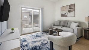A modern living room with a sofa, a coffee table, a TV, and a sliding glass door leading to an outdoor area. - Landing | Beautiful 2BD, Pool (Casas Adobes)