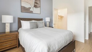 A neatly made bed with white bedding and two pillows, a wooden nightstand with a lamp, and a wall-mounted abstract painting. - Landing | Incredible 2BD, Pool (Casas Adobes)