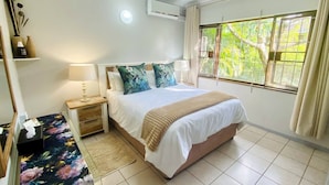 A bedroom with a bed, bedside tables, a nightstand, a window with curtains, and an air conditioning unit.