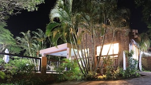 A brick building with a covered entrance, surrounded by lush greenery and palm trees.