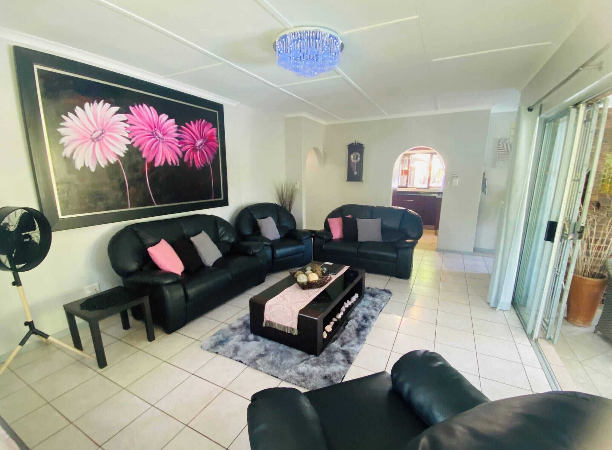 A living room with a black sofa set, a coffee table, a large floral painting, a chandelier, and a view into the kitchen.