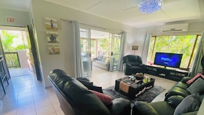 A living room with a black leather sofa, a glass coffee table, a TV, and a balcony with a view of greenery.