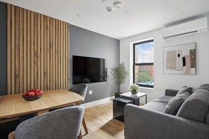 A modern living room with a wooden panel wall, a flat-screen TV, a dining table with a bowl of fruit, grey upholstered furniture, and a window with a view of buildings. - 1 Bedroom New Renovated - Wash/Dryer & Kitchenette -G (New York)