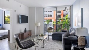 A modern living room with a sofa, armchair, coffee table, TV, and a balcony with a view of buildings and greenery. - Landing | Chic Studio, Yoga Studio, Game Room (Miami)